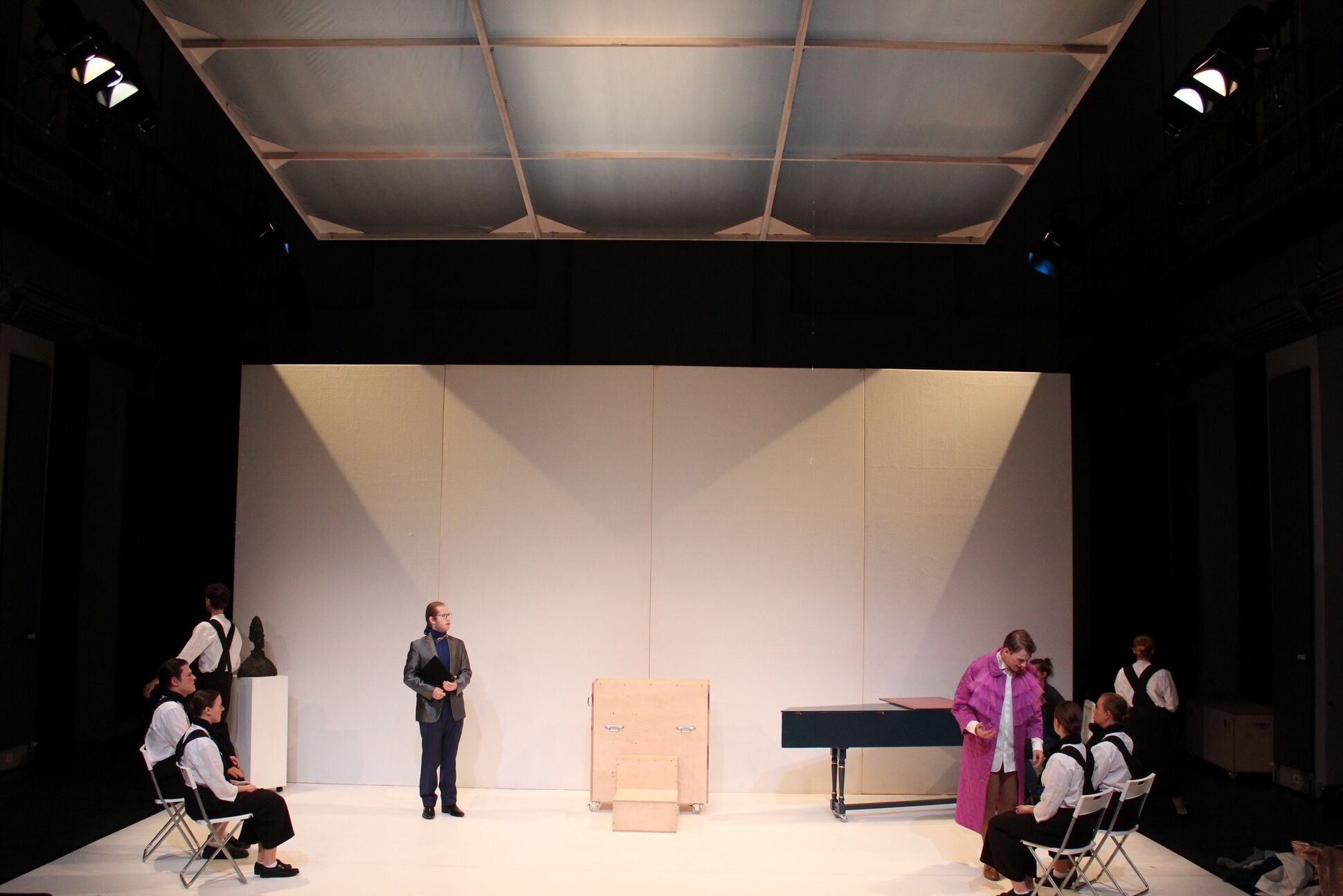 Bühnenbild: Auf der weißen Bühne sind Menschen und verschiedene Objekte zu sehen.